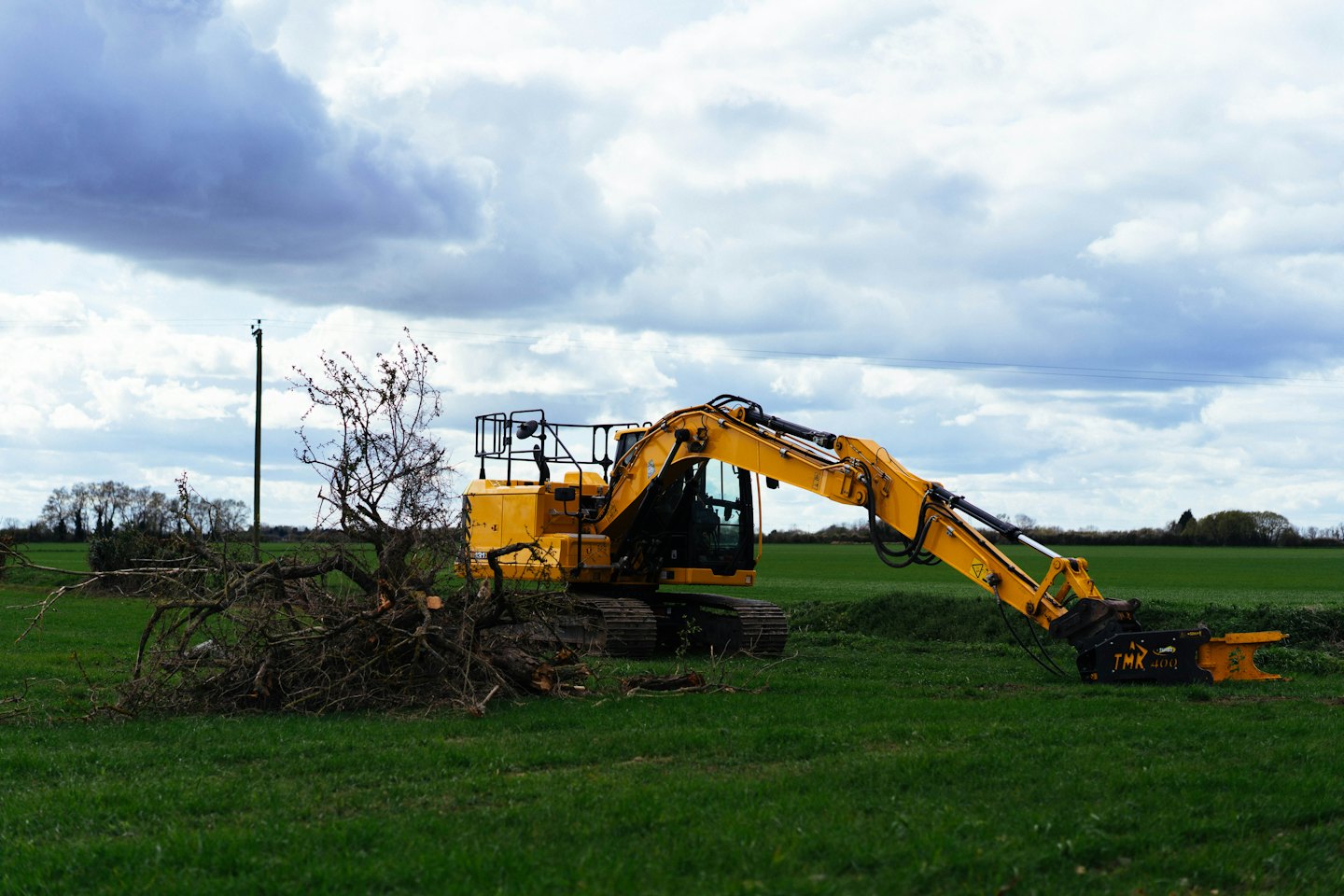 A yellow excavator with a tree shear clears branches in a field.