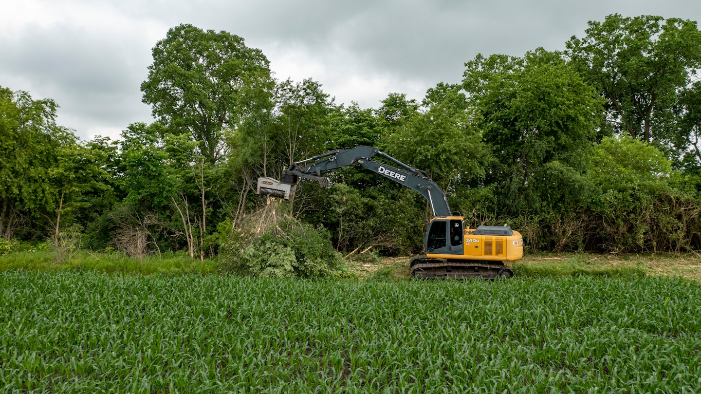 Yellow excavator mulching dense brush in a rural field.